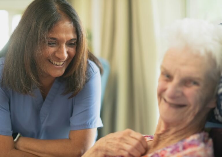 A smiling older adult sits in a cozy living room while a caregiver helps with daily activities nearby.