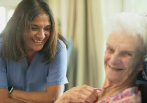 An older adult and caregiver share tea and conversation at a bright kitchen table.